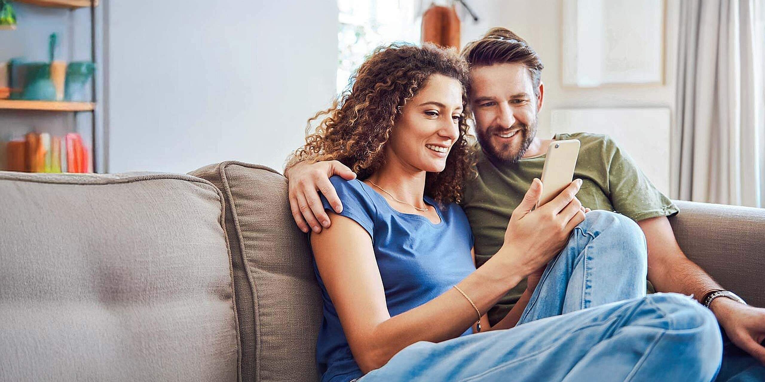 Ein lächelndes Paar sitzt auf einem Sofa, der Mann hat den Arm um die Frau gelegt, beide schauen auf das Smartphone in der Hand der Frau. Sie wirken entspannt und zufrieden, umgeben von einem sanft beleuchteten, gemütlichen Raum mit großen Fenstern im Hintergrund.