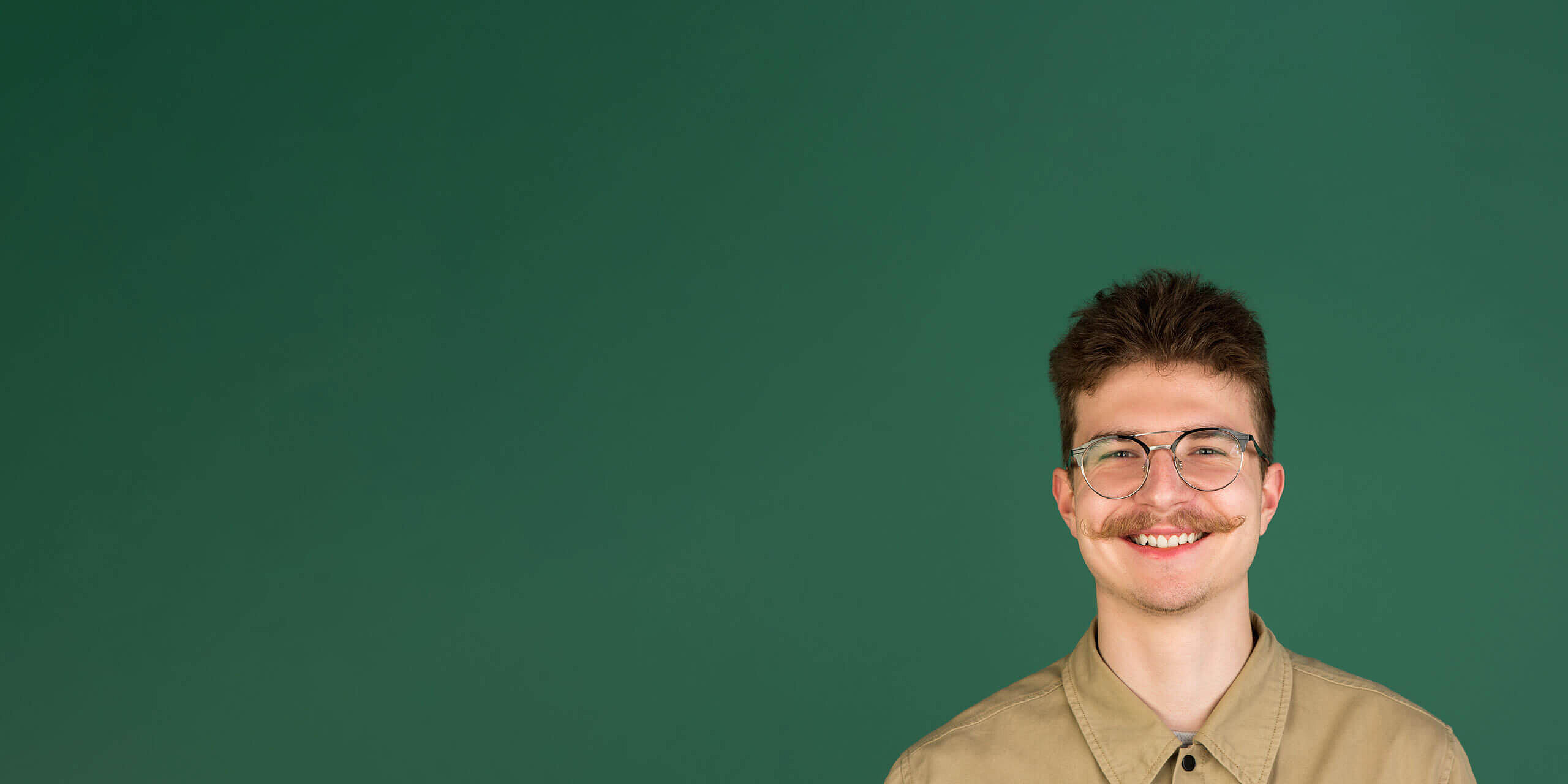 Eine Person mit kurzen dunklen Haaren, Brille und Schnurrbart lächelt in die Kamera. Sie trägt ein beiges Hemd mit Kragen. Der Hintergrund ist einfarbig grün.