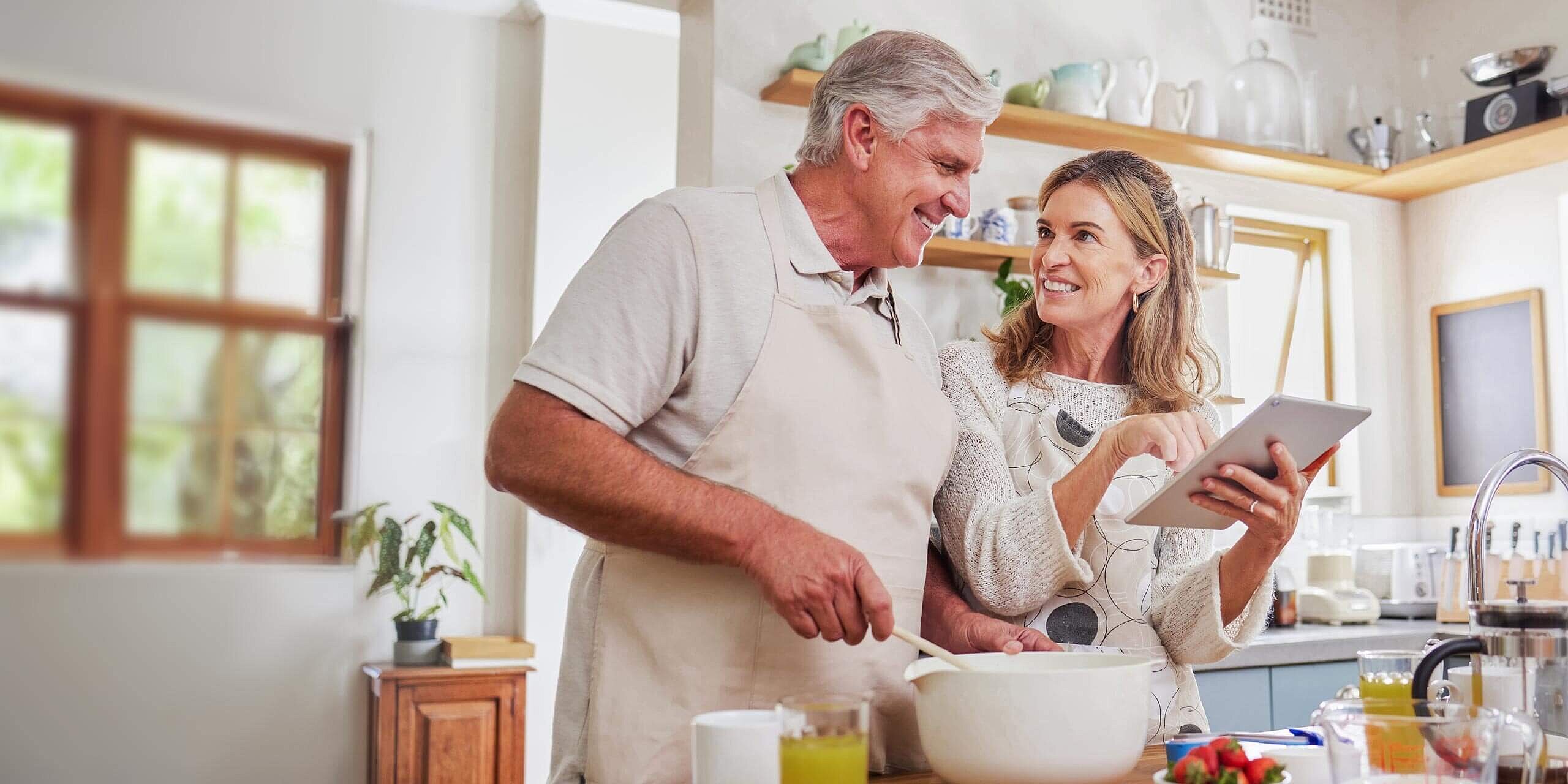 Ein lächelndes älteres Paar kocht gemeinsam in einer hellen Küche. Der Mann mischt Zutaten in einer Schüssel, während die Frau ein Tablet hält und darauf zeigt. Die Theke ist mit verschiedenen Küchenutensilien gefüllt, darunter Eier, Saft und Obst.