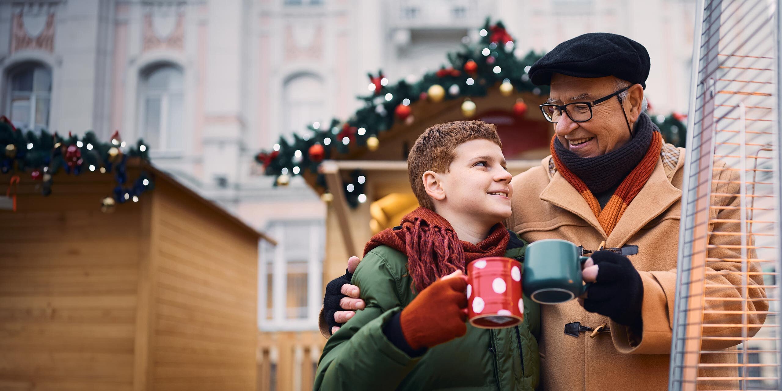 Großvater und Enkel genießen im Winter warme Getränke neben einem Außenheizstrahler. Sie sind gemütlich gekleidet und stehen vor festlich geschmückten Marktständen, die mit Girlanden und Ornamenten geschmückt sind.