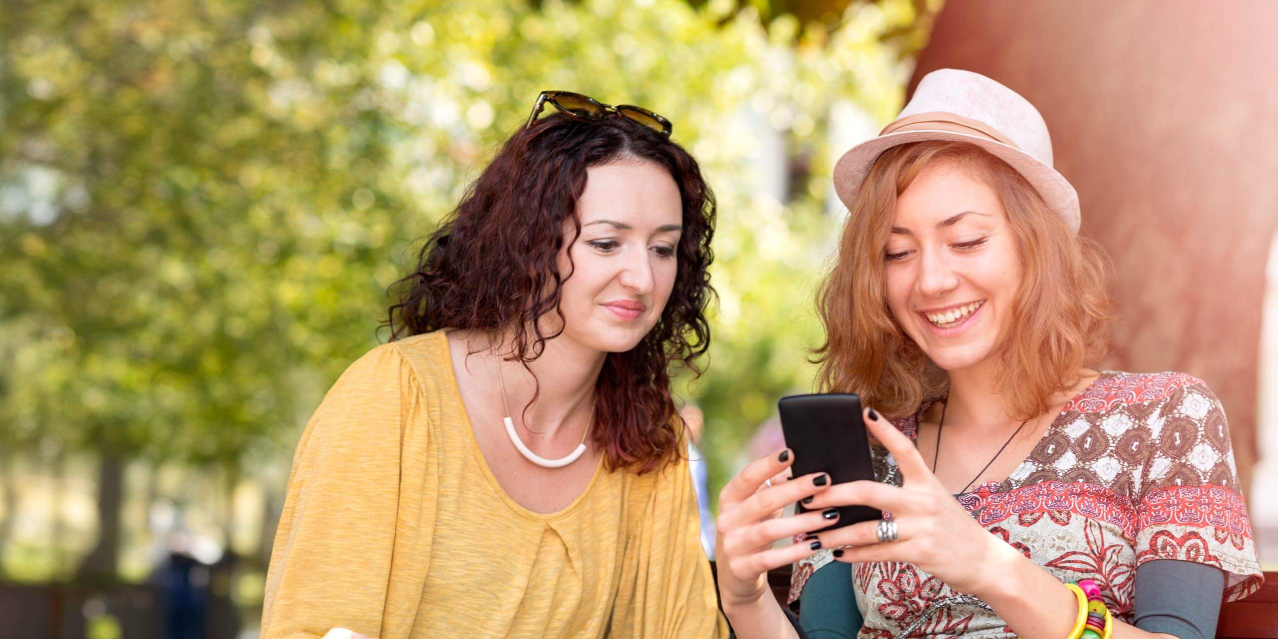 Zwei Frauen sitzen im Freien und lachen, während sie auf ein Smartphone schauen. Eine Frau mit lockigem dunklem Haar trägt ein gelbes Oberteil und eine Sonnenbrille auf dem Kopf. Die andere mit hellbraunem Haar und Hut trägt ein buntes Hemd und mehrere Ringe. Im Hintergrund sind Bäume und Sonnenlicht zu sehen.