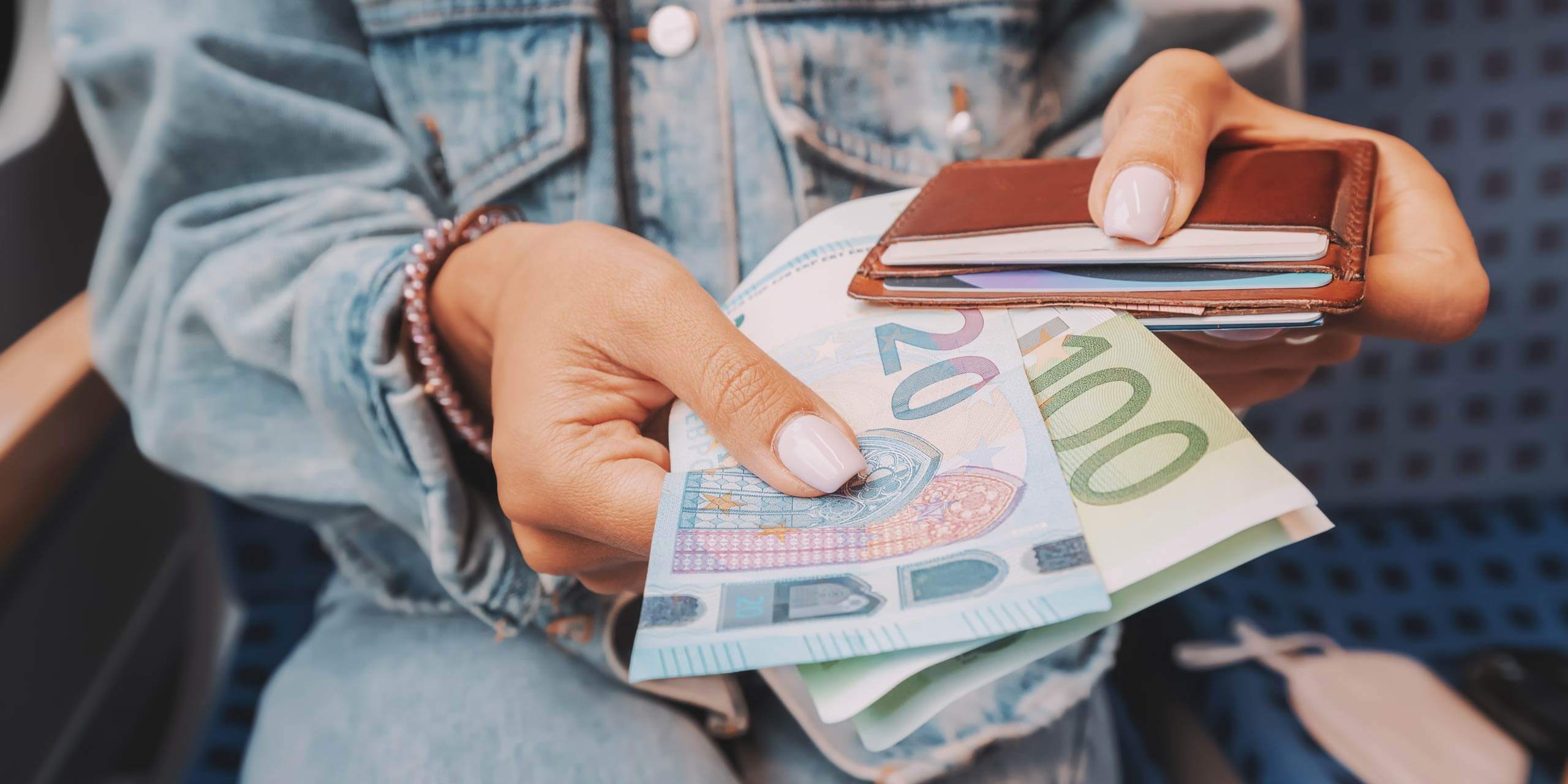 Eine Person in einer Jeansjacke hält in der einen Hand Euro-Banknoten (20 und 100) und in der anderen eine braune Geldbörse. Der Hintergrund ist leicht unscharf.