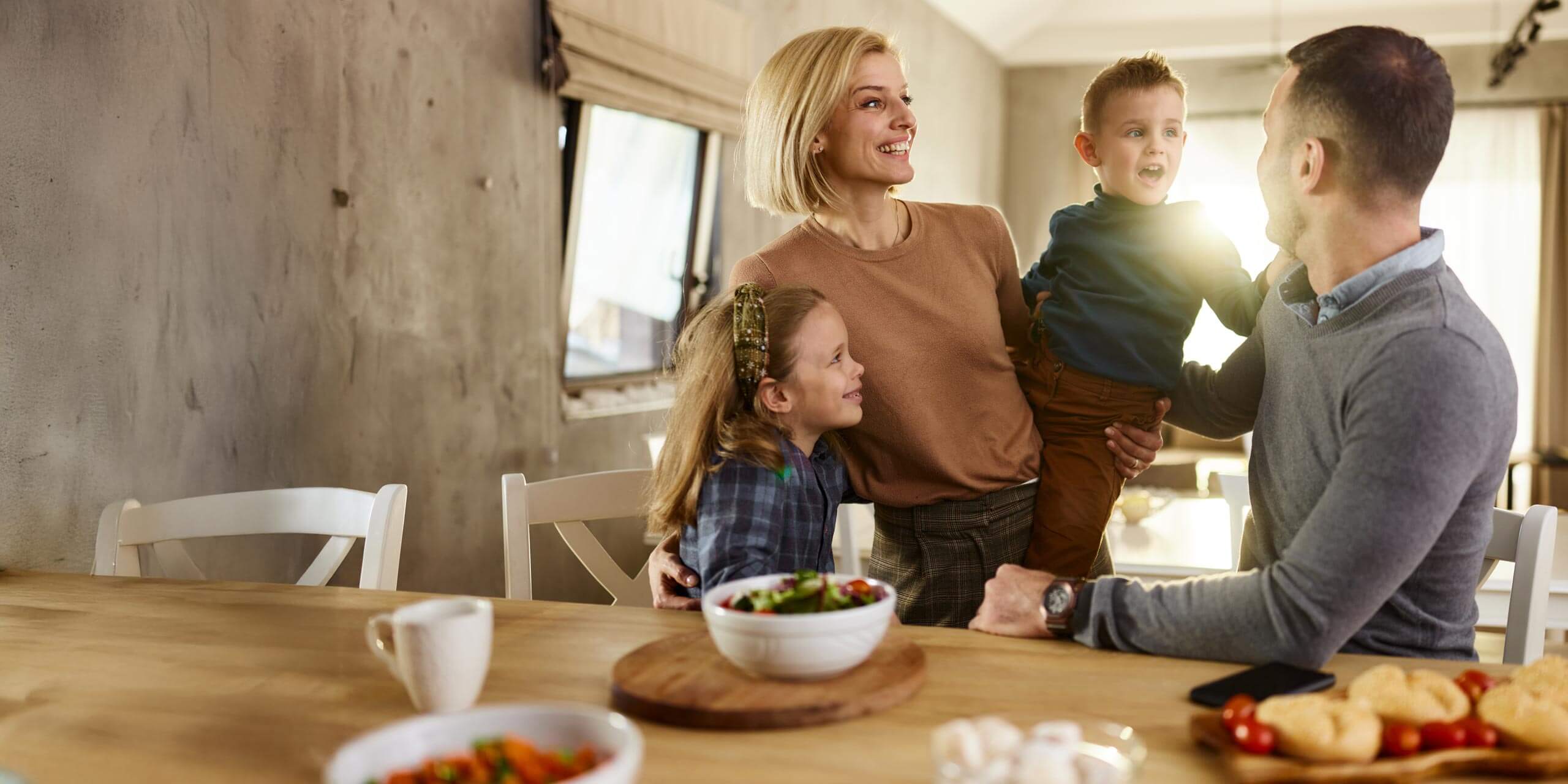 Eine vierköpfige Familie versammelt sich um einen Esstisch und hat Essen, darunter eine Schüssel Salat und Gebäck. Die Mutter hält einen kleinen Jungen im Arm, während Vater und Tochter daneben stehen. Alle lächeln und schauen sich herzlich an, während das Sonnenlicht durch das Fenster fällt.