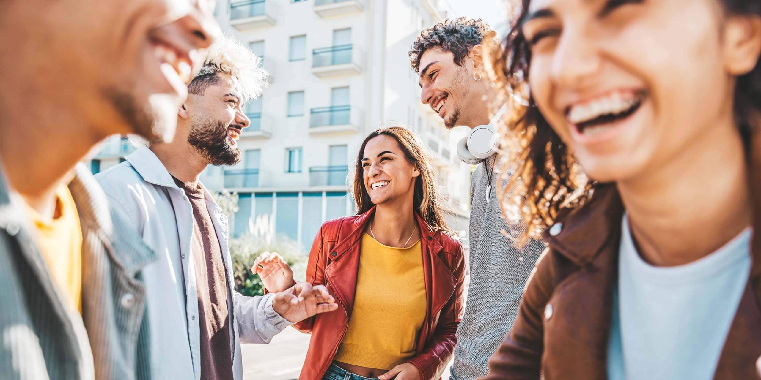 Eine Gruppe von fünf Personen lacht und lächelt im Freien. Sie scheinen Freunde zu sein, die gemeinsam einen schönen Tag verbringen. Eine Person trägt Kopfhörer um den Hals. Im Hintergrund ist ein modernes Gebäude mit Balkonen zu sehen. Die allgemeine Stimmung ist fröhlich und lebhaft.