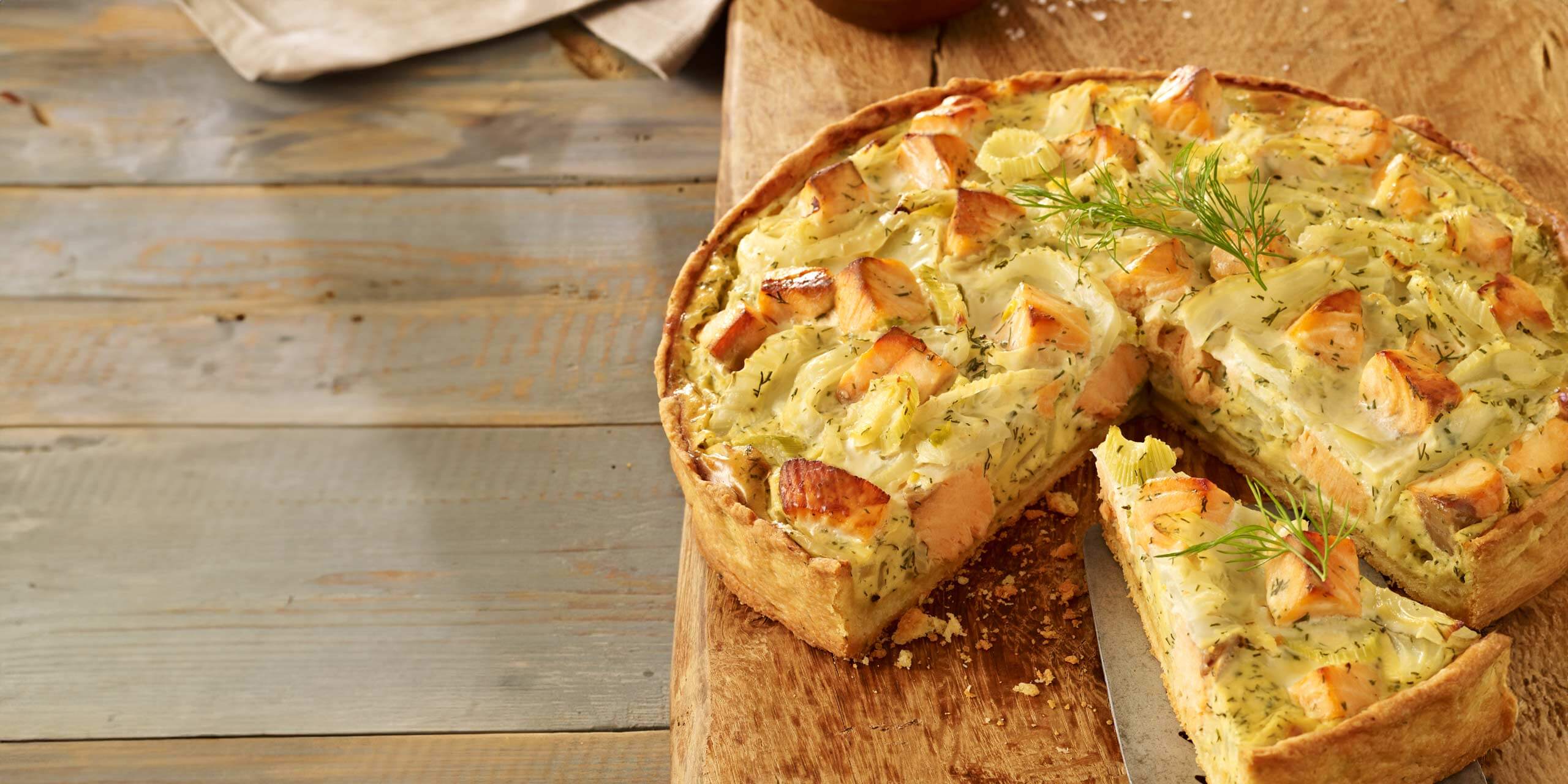 Eine rustikale Fechel-Lachs-Quiche, von der eine Scheibe entfernt wurde, präsentiert ihre Füllung aus gewürfeltem Gemüse, Kräutern und einer cremigen Mischung. Die goldene Kruste und der hellbraune Belag werden auf einem Holzschneidebrett serviert, und etwas frischer Dill garniert die Quiche.